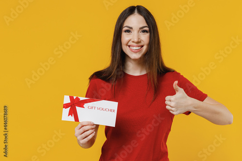 Smiling excited fun young brunette woman 20s wears basic red t-shirt hold gift certificate coupon voucher card for store showing thumb up like gesture isolated on yellow background studio portrait.