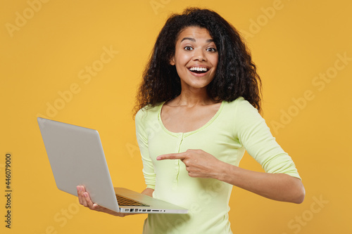 Satisfied happy smiling jubilant cheerful african american young brunette woman 20s wears green shirt hold use work on laptop pc computer pointing on it isolated on yellow background studio portrait. © ViDi Studio