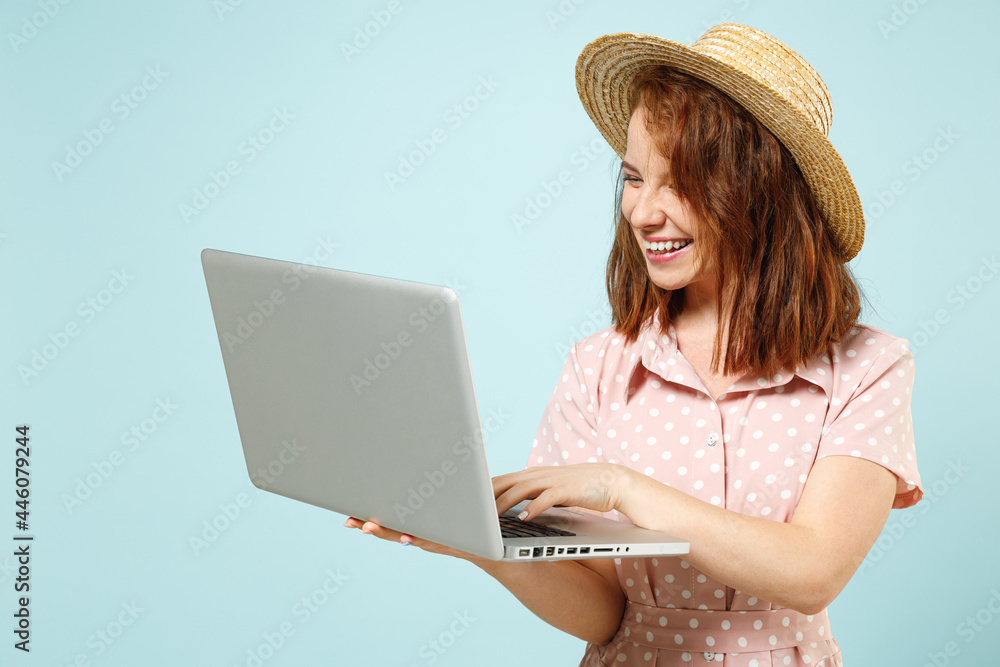 Smiling busy happy young redhead curly woman 20s wear casual pink dress straw hat hold use work on laptop pc computer typing hands on keyboard isolated on pastel blue color background studio portrait