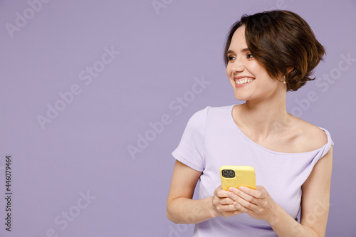Young smiling happy woman 20s with bob haircut in white t-shirt using mobile cell phone chat online browsing internet look aside on workspace area isolated on pastel purple background studio portrait.