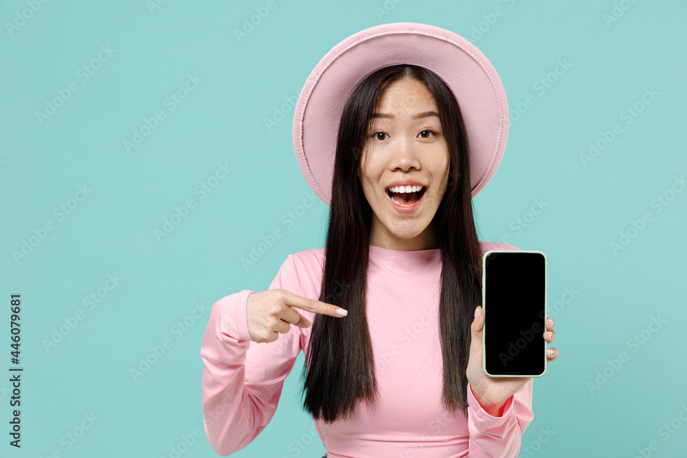 Astonished happy young brunette asian woman 20s wears pink clothes hold in hand mobile cell phone with blank screen workspace area point on it isolated on pastel blue color background studio portrait Astonished happy young brunette asian woman 20s wears pink clothes hold in hand mobile cell phone with blank screen workspace area point on it isolated on pastel blue color background studio portrait