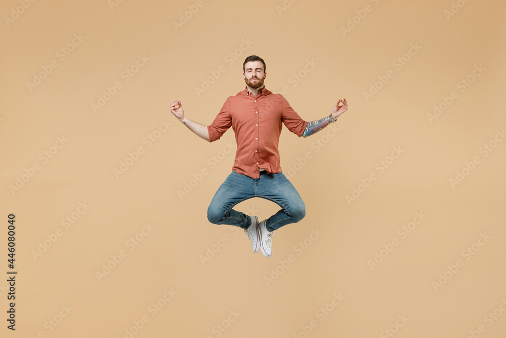 Full size body length tatooed young brunet man 20s short haircut wears apricot shirt jump hold spread hands in yoga om aum gesture relax meditate isolated on pastel orange background studio portrait.