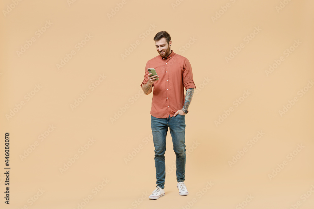 Full size body length smiling cheerful tatooed young brunet man 20s short haircut wears apricot shirt go stride hold in hand use mobile cell phone isolated on pastel orange background studio portrait Full size body length smiling cheerful tatooed young brunet man 20s short haircut wears apricot shirt go stride hold in hand use mobile cell phone isolated on pastel orange background studio portrait