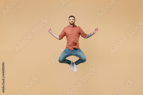 Full size body length tatooed young brunet man 20s short haircut wears apricot shirt jump hold spread hands in yoga om aum gesture relax meditate isolated on pastel orange background studio portrait. © ViDi Studio