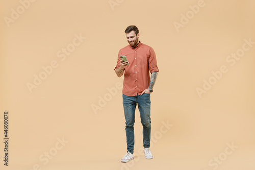 Full size body length smiling cheerful tatooed young brunet man 20s short haircut wears apricot shirt go stride hold in hand use mobile cell phone isolated on pastel orange background studio portrait