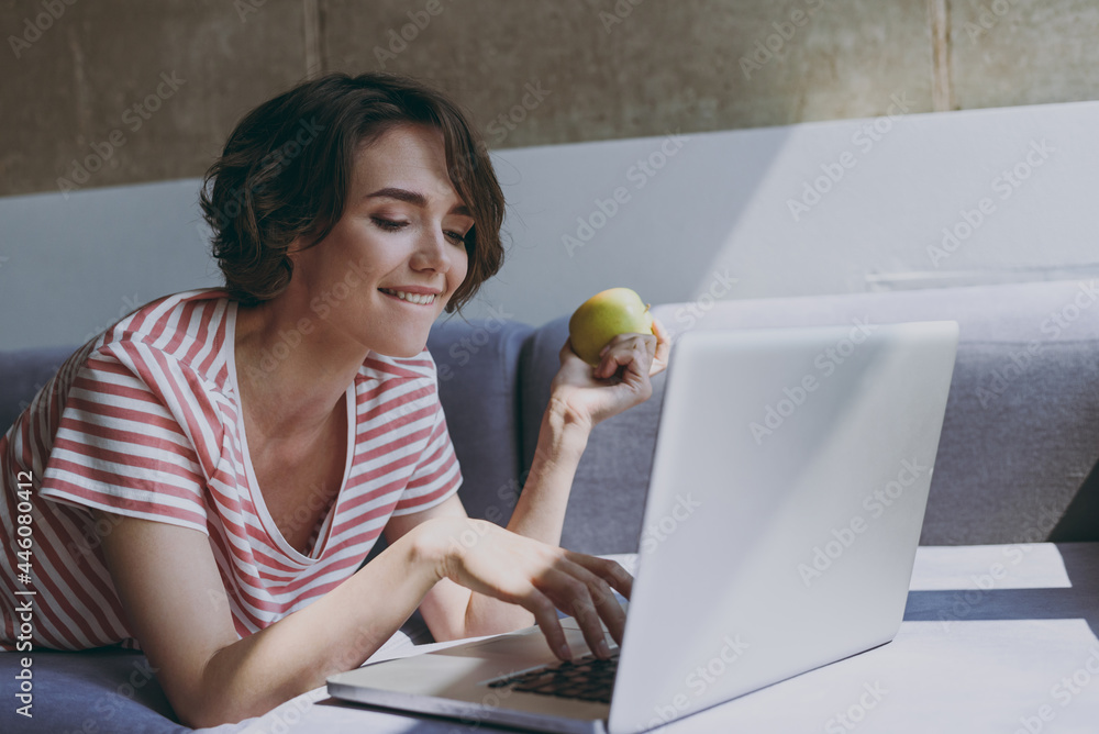 Young vegeterian woman in casual clothes lying on grey sofa work online with laptop pc computer typing message eat green apple fruit rest relax indoors flat at home People lifestyle leisure concept Young vegeterian woman in casual clothes lying on grey sofa work online with laptop pc computer typing message eat green apple fruit rest relax indoors flat at home People lifestyle leisure concept