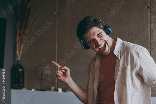 Young overjoyed satisfied cheerful happy caucasian positive man wear casual clothes beige shirt pink t-shirt headphones dancing listen music rest indoors at home on weekends on brown wall background