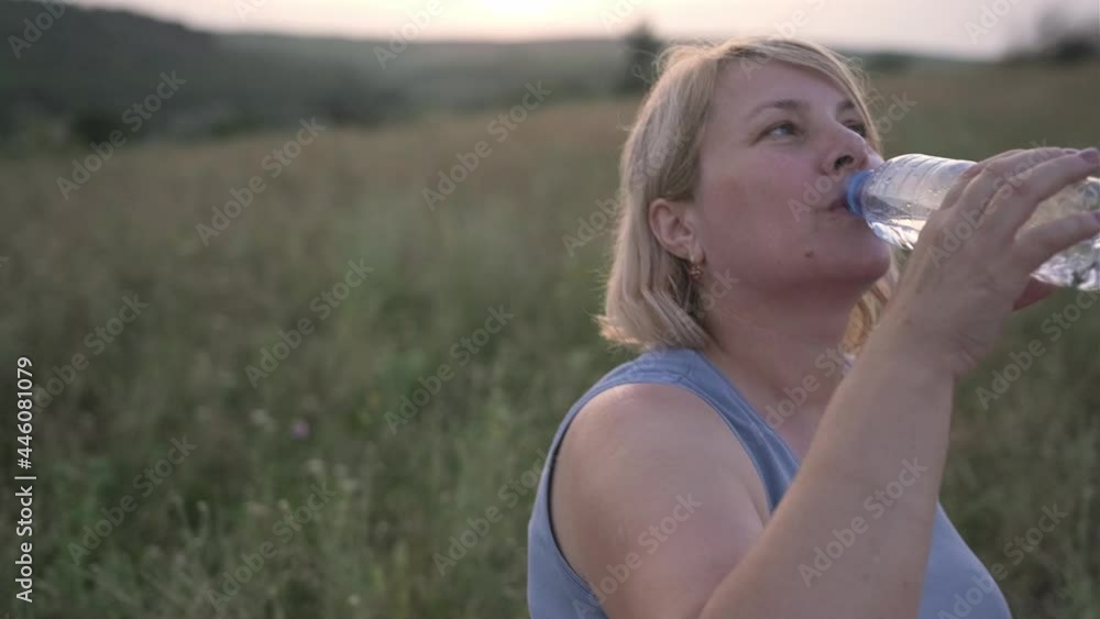 Woman in sports wear sitting in the middle of a field on a fitness carpet and drinking some water after exercises. Woman in sports wear sitting in the middle of a field on a fitness carpet and drinking some water after exercises.