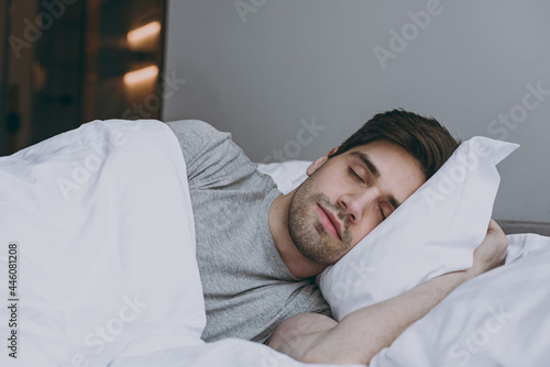 Calm brunet tranquil european young man 20s wearing pajamas grey t-shirt lying in bed sleep slumber resting with closed eyes relaxing at home indoors bedroom. Good mood night morning bedtime concept