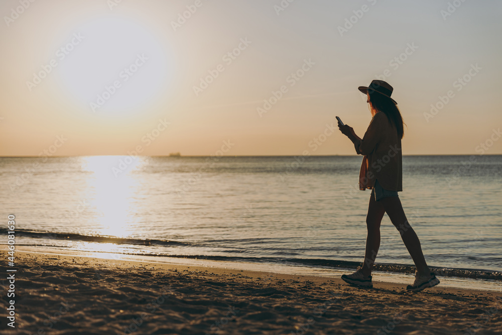 Side view full length young woman wearing straw hat shirt summer clothes walk use modile cell phone outdoors at sunrise sun dawn over sea beach background. People vacation lifestyle journey concept