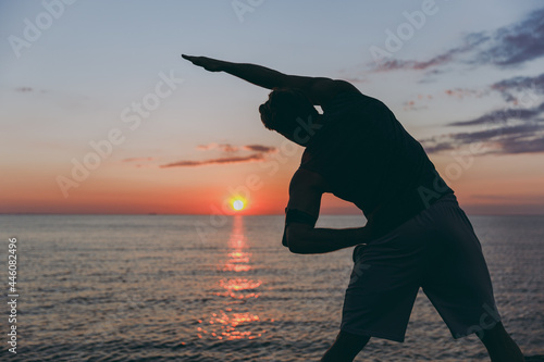 Silhouette back view young strong athletic toned fit sportsman man in sports clothes warm up training do side bends at sunrise sun dawn over sea beach outdoor on pier seaside in summer day morning