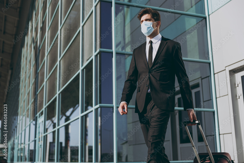Bottom view young traveler european businessman man 20s wear black dinner suit facial mask walking go outside at international airport terminal with suitcase valise Air flight business trip concept.