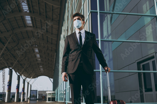Bottom view young caucasian traveler businessman man in black dinner suit sterile facial mask walk go outside at international airport terminal with suitcase valise Air flight business trip concept