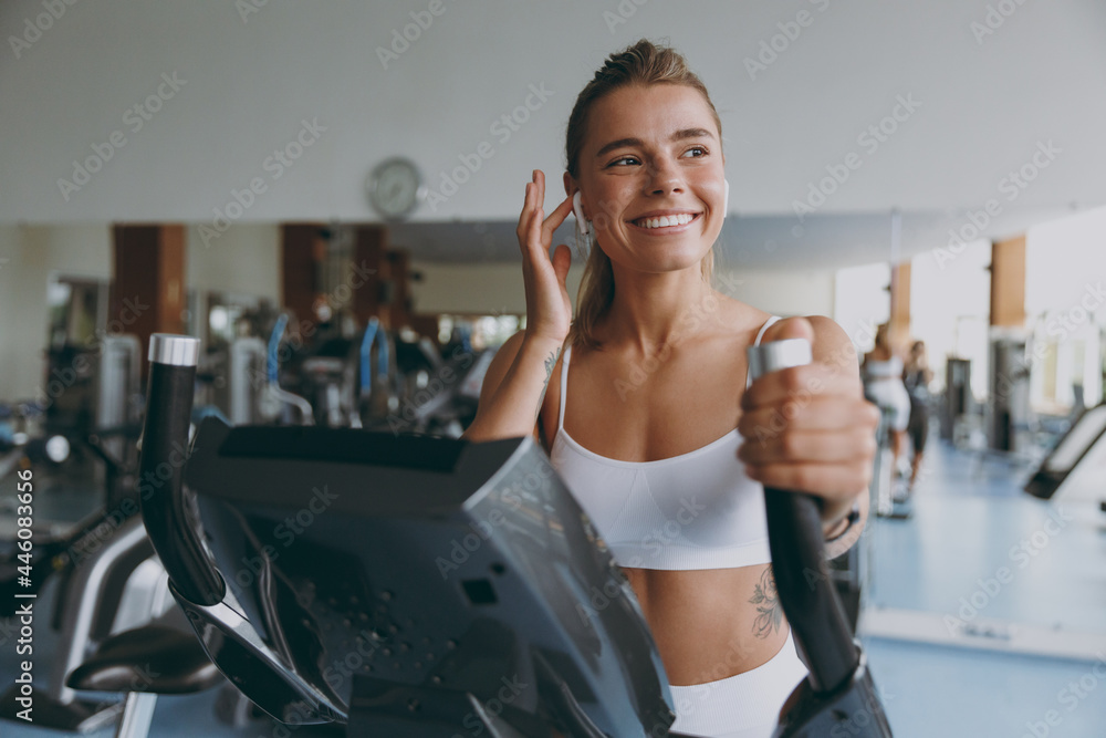 Front view smiling satisfied young strong sporty athletic sportswoman woman in white sportswear earphones listen music look aside warm up train use exercise bike in gym indoor Workout sport concept Front view smiling satisfied young strong sporty athletic sportswoman woman in white sportswear earphones listen music look aside warm up train use exercise bike in gym indoor Workout sport concept