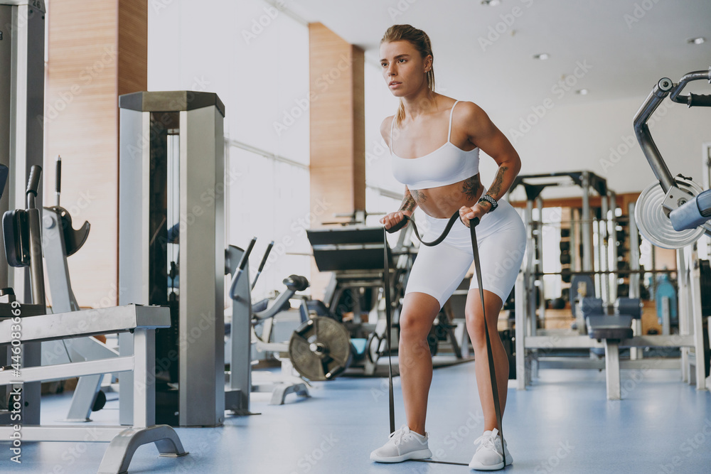 Full size young skinny strong sporty athletic sportswoman woman 20s wearing white sportswear warm up training pull using fitness rubber bands in gym indoor Workout sport motivation lifestyle concept Full size young skinny strong sporty athletic sportswoman woman 20s wearing white sportswear warm up training pull using fitness rubber bands in gym indoor Workout sport motivation lifestyle concept