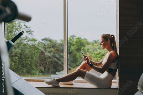 Full body young side profile view sporty woman in white sportswear earphones listen music use mobile phone look at smart watch siton windowsill of gym near treadmill rest indoor People sport concept photo