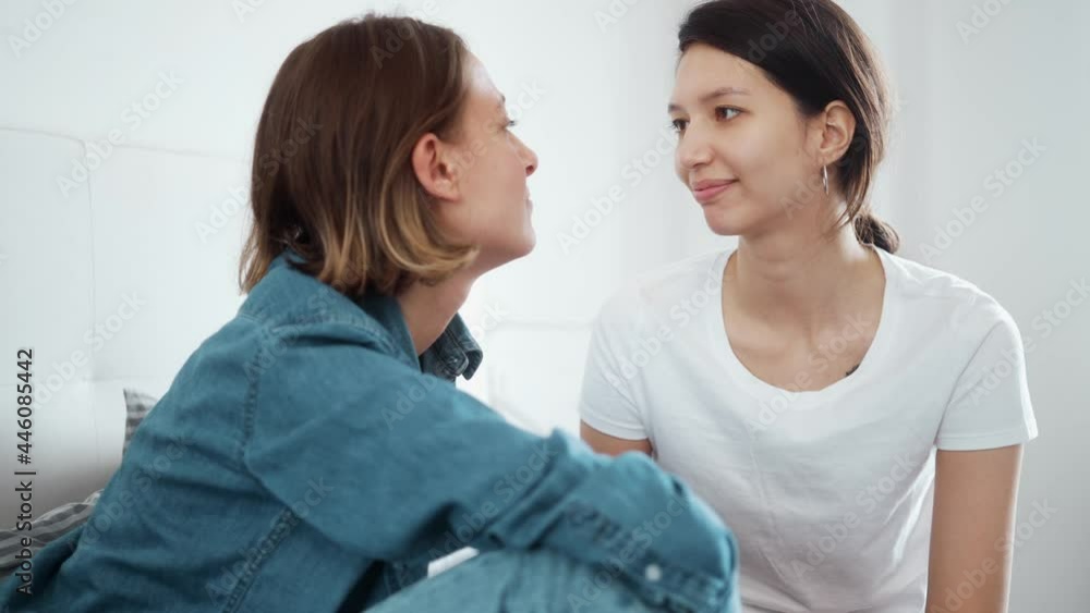 A smiling lesbian couple are looking at each other while kissing and hugging sitting together at the bedroom A smiling lesbian couple are looking at each other while kissing and hugging sitting together at the bedroom