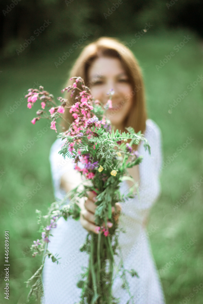 A beautiful happy girl in a white dress holds out a bouquet of wild flowers to the camera and smiles. Portrait of a slender girl with a bouquet of wild flowers in nature. Selective focus