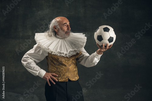 Elderly gray-haired man, actor posing with football ball isolated on dark vintage background. Retro style, comparison of eras concept.