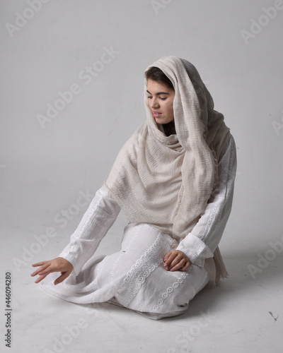 Fotografie Full length portrait of young woman wearing classical white gown and a head covering veil in biblical style, sitting pose on light studio background