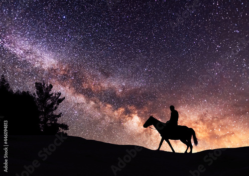 Silhouette of a lone rider on a horse standing on a hill in the starry night. Behind him is the beautiful bright Milky Way galaxy.