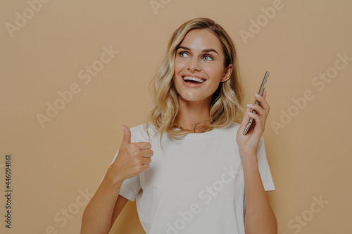 Young satisfied girl in casual clothes holding mobile phone and showing thumb up gesture, being excited