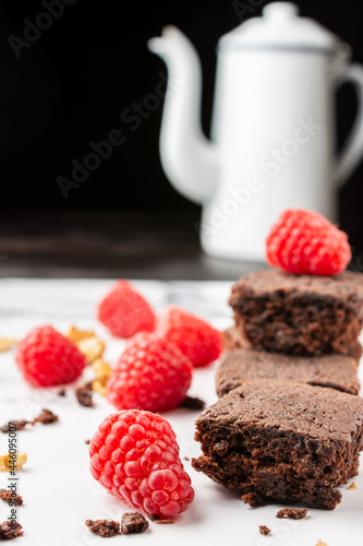 Close-up of chocolate brownie with crumbs  walnuts  red raspberries and white coffee pot  selective focus  on white marble table  black background  vertical