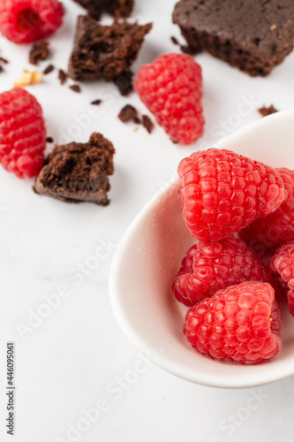 Top view of white bowl with raspberries, pieces of chocolate brownie and walnuts, selective focus, on white table, vertical, with copy space