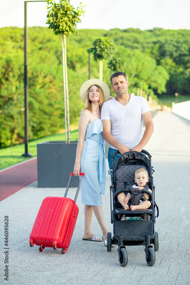 happy young parents mom and dad with a red suitcase and a baby in a stroller are going on a vacation trip in the summer, smiling happy young parents mom and dad with a red suitcase and a baby in a stroller are going on a vacation trip in the summer, smiling