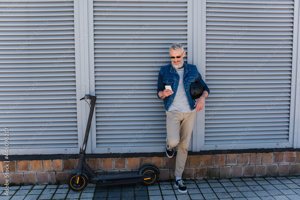 full length of happy and mature man in sunglasses using smartphone near e-scooter