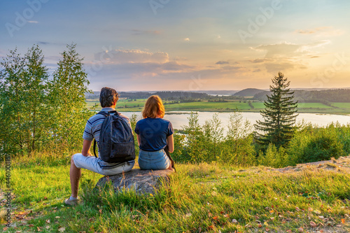 Tableau sur toile Rear view of couple sitting on hill against beautiful summer landscape with forest and lake in summer evening at sunset