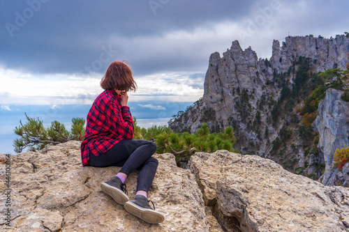 Rear view of young woman sitting on mountain cliff stones and admiring beautiful landscape of mountainous area and sky. Teen tourist girl against clouds in summer day. Travel destinations, staycation