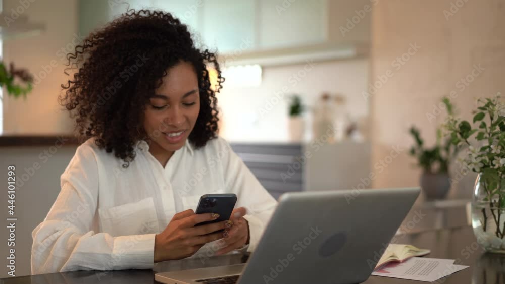 Joyful young mixed race woman chatting use smartphone enjoying break during work in front of laptop. Smiling female typing message or texting on mobile sitting on desk at home office Joyful young mixed race woman chatting use smartphone enjoying break during work in front of laptop. Smiling female typing message or texting on mobile sitting on desk at home office