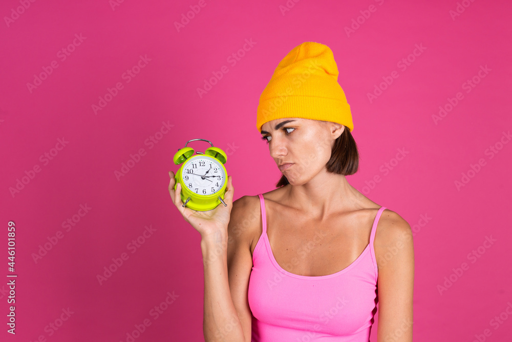 Bright portrait of woman in yellow hat on pink  background with alarm clock angry tired look at watch