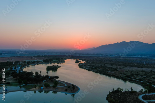 a stunning aerial shot of a sunset over the lake surrounded majestic mountain ranges and lush green trees and grass at Santa Fe Dam Recreation Area in Irwindale, California photo