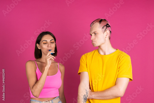 Bright portrait of young beautiful couple on pink background smoking electronic cigarette, woman enjoying smiling,man looks unhappy disappointed