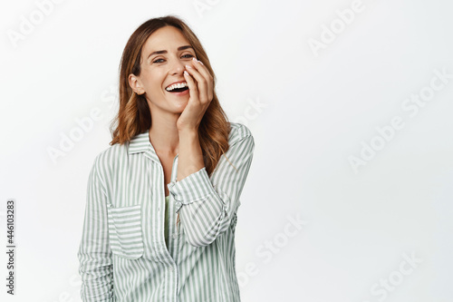 Carefree happy woman in blouse, laughing and smiling cheerful, touching face and looking upbeat at camera, standing against white background