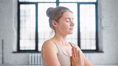 Pretty woman with closed eyes in beige sportswear breathes deeply holding hands in namaste mudra at chest in yoga studio