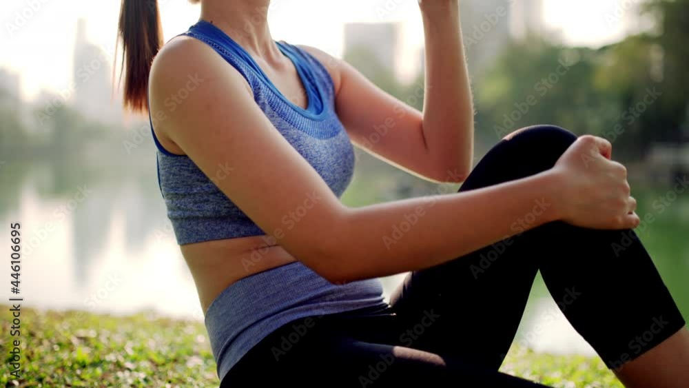 A woman is sitting on the grass and drinking water from a bottle beside a pond in the city park A woman is sitting on the grass and drinking water from a bottle beside a pond in the city park