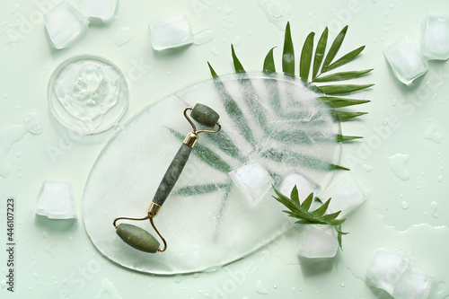Fototapeta Naklejka Na Ścianę i Meble -  Self made moisturizer and green jade face roller on oval piece of ice and ice cubes. Exotic palm leaves on mint green background. Minimal flat lay, top view. Facial massage, handmade cosmetics.