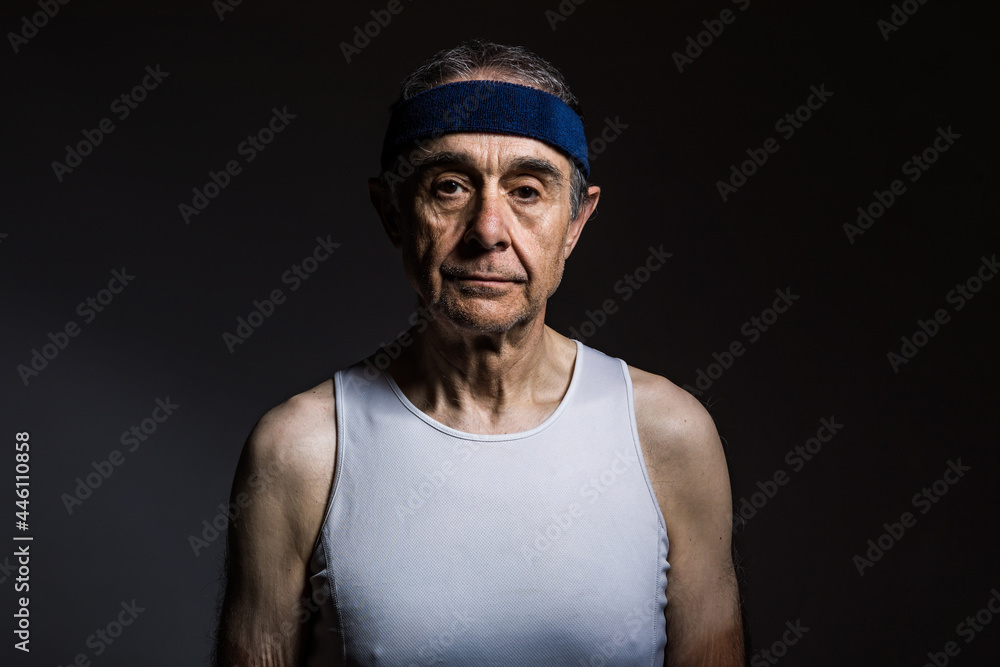 Elderly athlete wearing white sleeveless shirt, with sun marks on the arms, and blue headband, on a dark background. Sports and victory concept. Elderly athlete wearing white sleeveless shirt, with sun marks on the arms, and blue headband, on a dark background. Sports and victory concept.