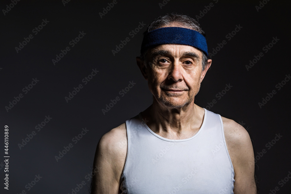 Fototapeta premium Elderly athlete wearing white sleeveless shirt, with sun marks on the arms, and blue headband, on a dark background. Sports and victory concept.