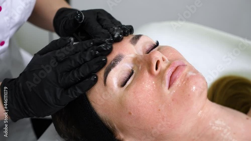 Hands in black nitrile gloves applying mask and massaging face of woman lying with eyes closed on couch. Closeup beautician performing rejuvenation procedure in wellness spa. Concept of skin care photo