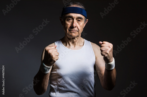 Elderly athlete wearing white sleeveless shirt, with sun marks on the arms, and blue headband clenching his fists, on a dark background. Sports and victory concept.