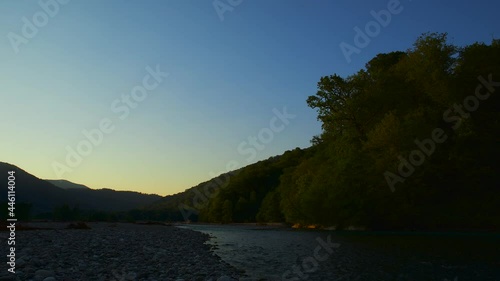 Dawn over a river in a wide mountain valley. The rays of the morning sun are increasingly illuminating the forest on the slopes of the mountains. The atmosphere of an early morning on the journey.