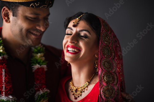 blurred indian man in turban looking at happy bride in traditional headscarf isolated on grey