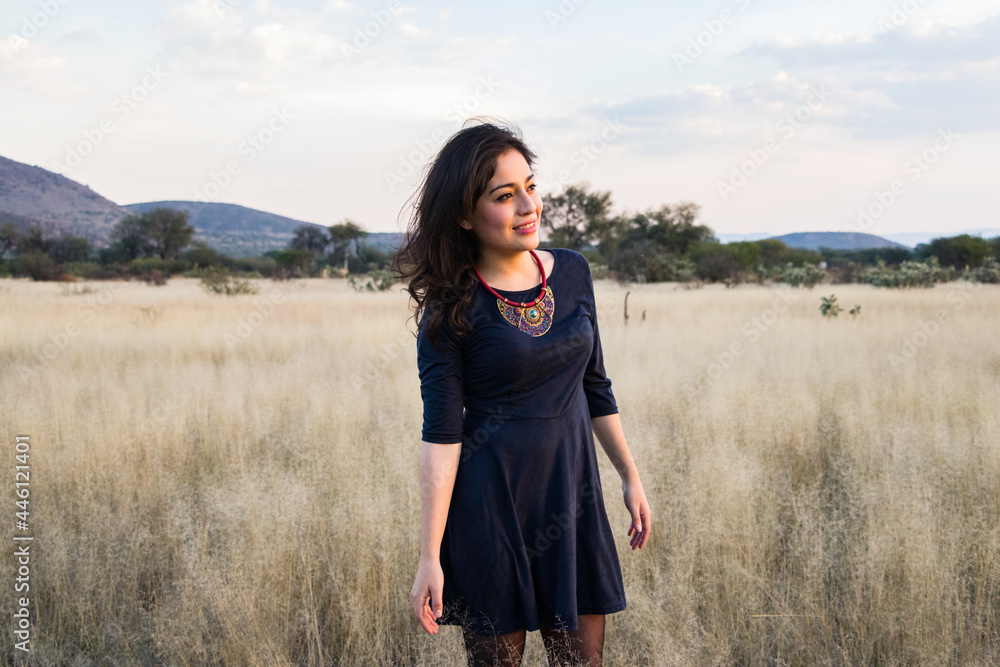 Young adult smiling Mexican woman standing in a dry grassy meadow looking into the distance