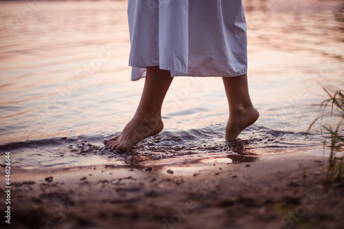Women's feet in the river. Lifestyle close-up on the feet of a woman in a long white dress walking along the riverbank at sunset.  © Юля Бурмистрова