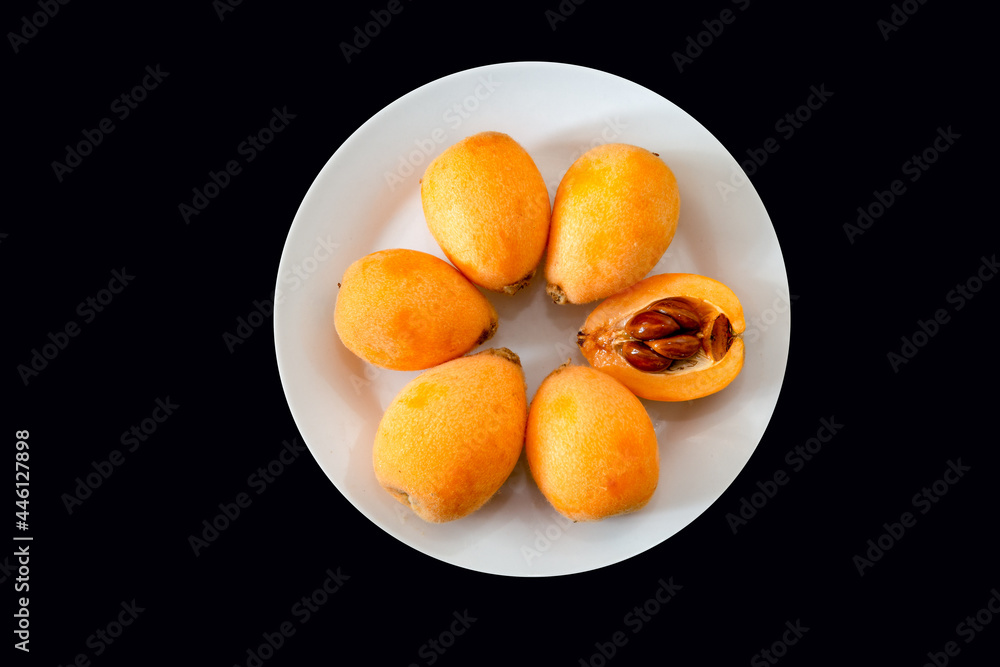 Ripe loquats on white plate on black background top view. One cut in half showing the inside of the fruit with the seeds ( Eriobotrya japonica )