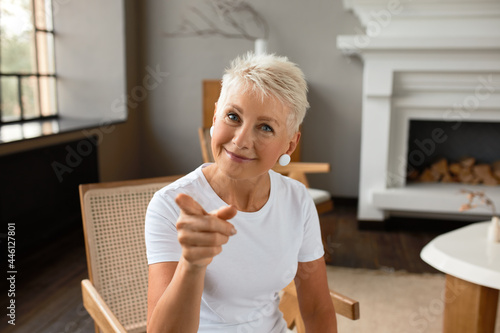 Cheerful mature female sit in living room pointing at you with finger. Ask question are you happy person. Concept of happiness, strong emotion, joy and appreciation of life concept